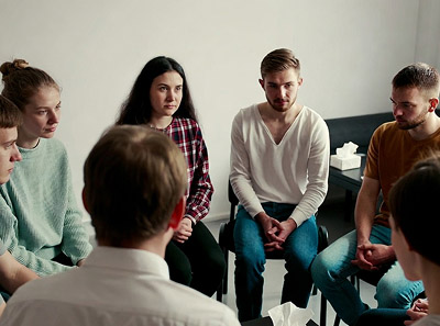 Группа молодых людей сидит в кругу на стульях в светлом помещении, внимательно слушают друг друга 
